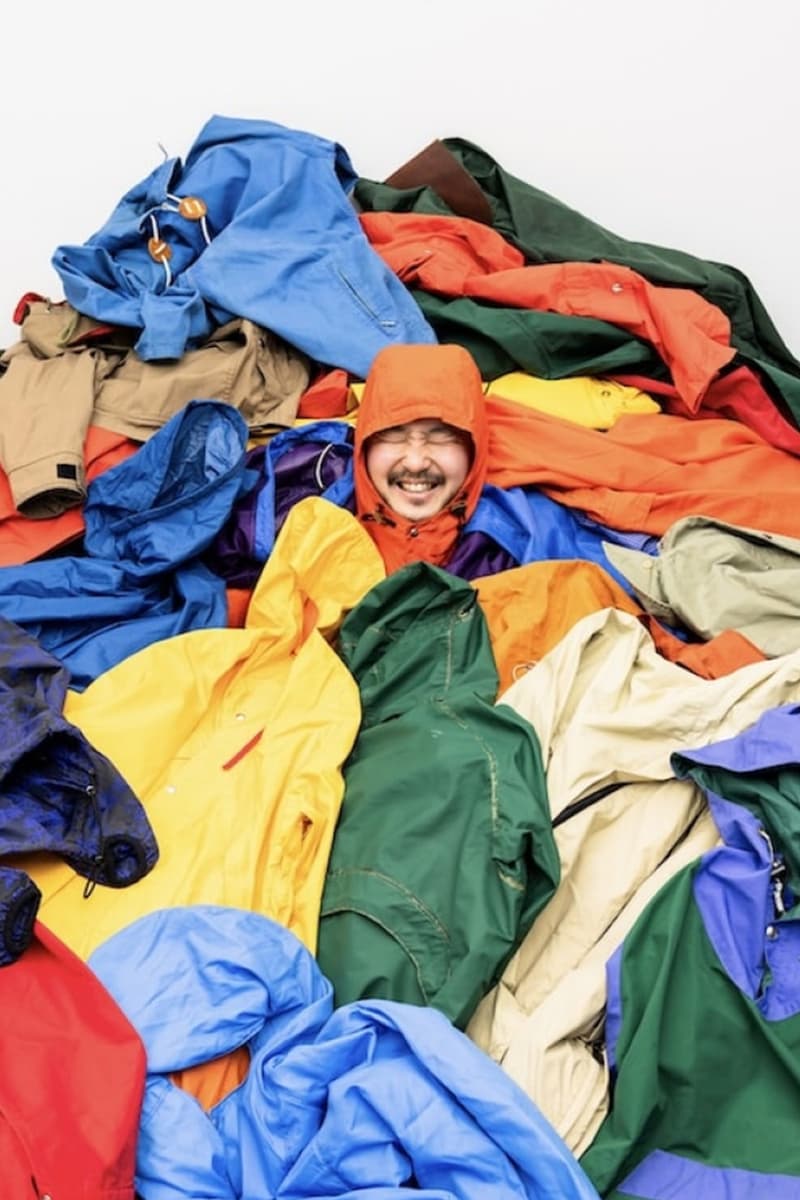 Personne souriante parmi des vestes colorées empilées, portant un manteau orange.