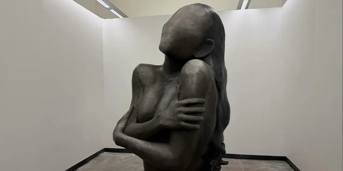 Sculpture sans visage, bras croisés, dans une galerie.
