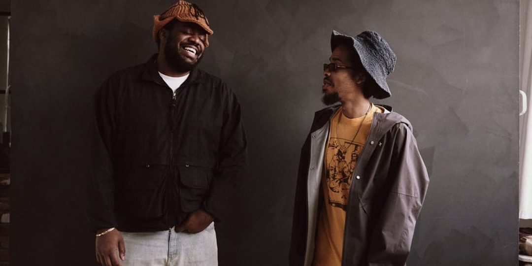 Deux hommes souriants portant des chapeaux et vestes devant un fond sombre.