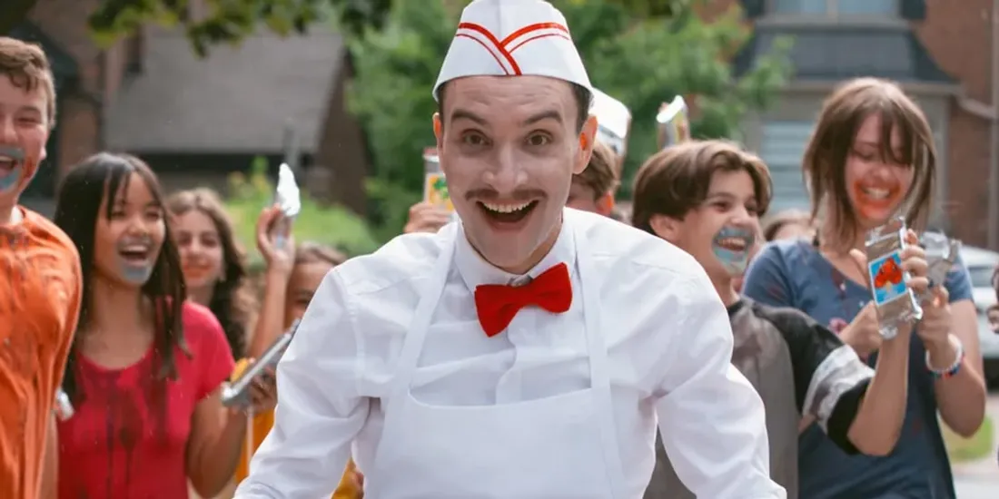 Homme avec moustache, chapeau de serveur, suivi par des enfants joyeux.