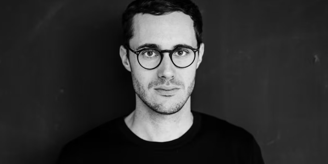 Homme aux lunettes et chemise noire sur fond sombre.