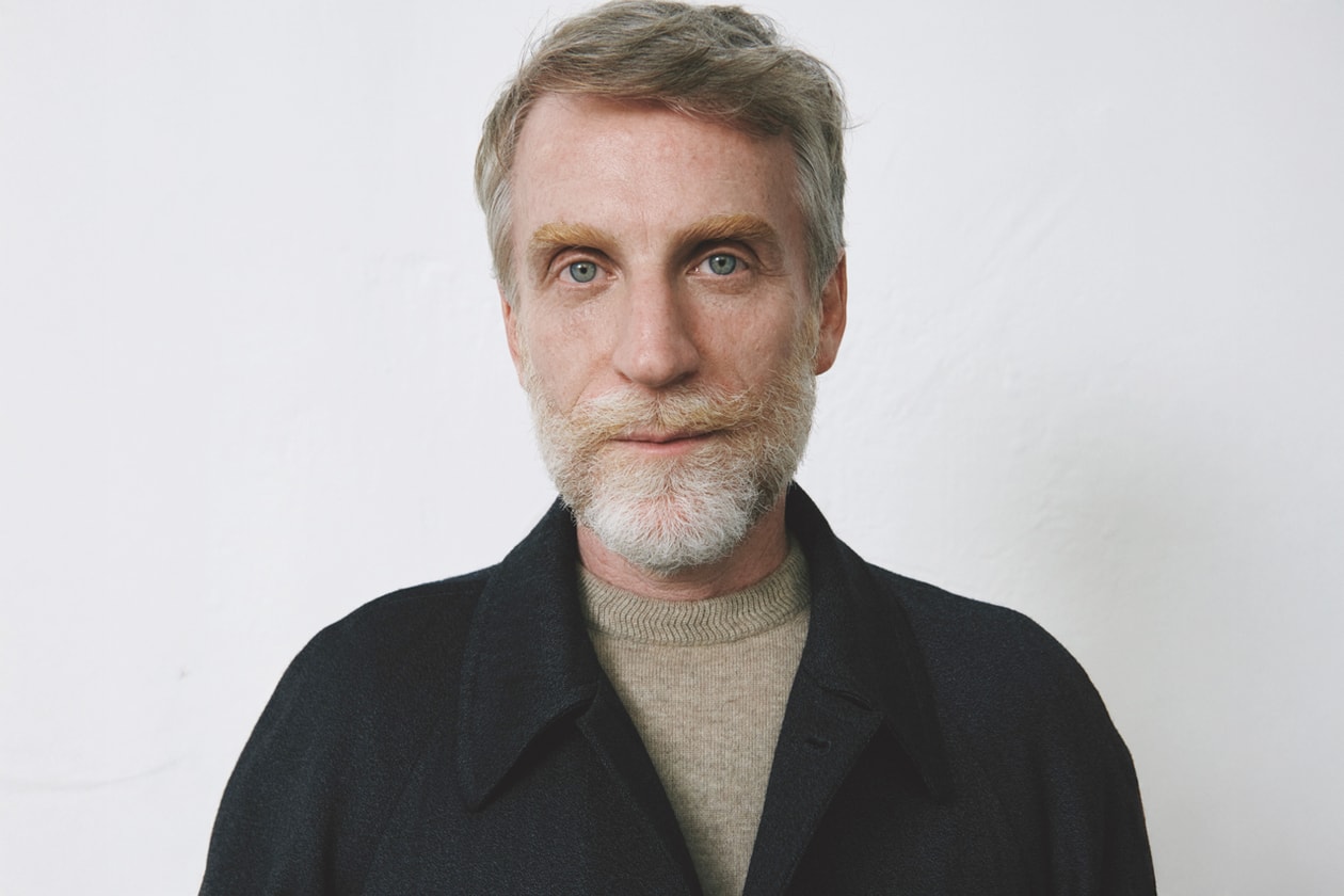 Homme aux cheveux gris et barbe posant sur un fond blanc.