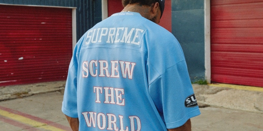 Homme portant un maillot bleu avec 'Screw the World' et 'Supreme' en arrière-plan urbain.