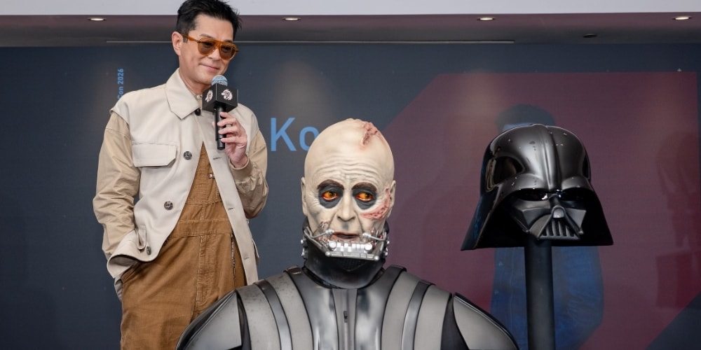 Homme avec des lunettes debout près de figures de science-fiction, casque noir visible.