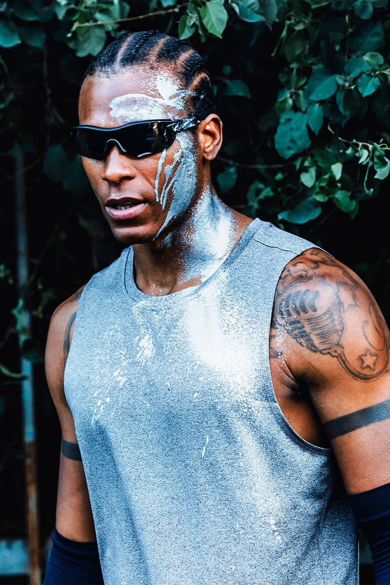 Homme avec lunettes de soleil, cheveux nattés, bras tatoués, T-shirt gris.