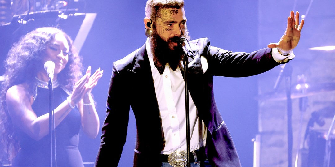 Homme barbu avec tatouages sur scène, microphone à la main.