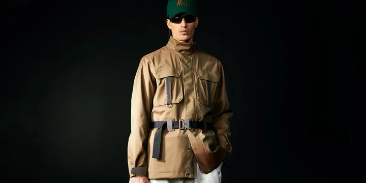 Homme en veste marron avec casquette verte et lunettes noires, fond noir.