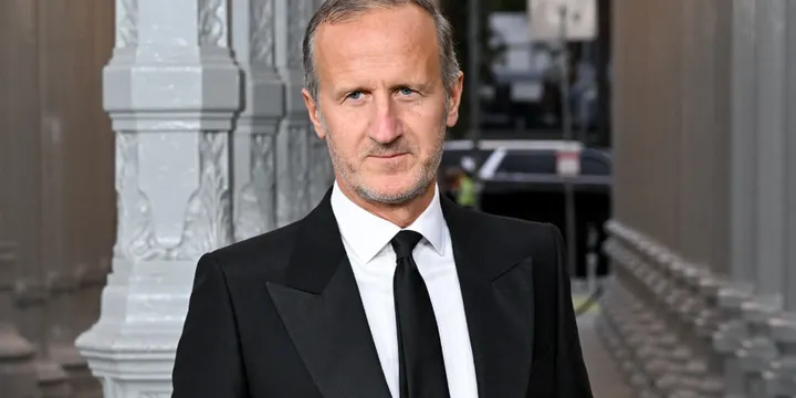 Homme en costume noir devant une architecture métallique.