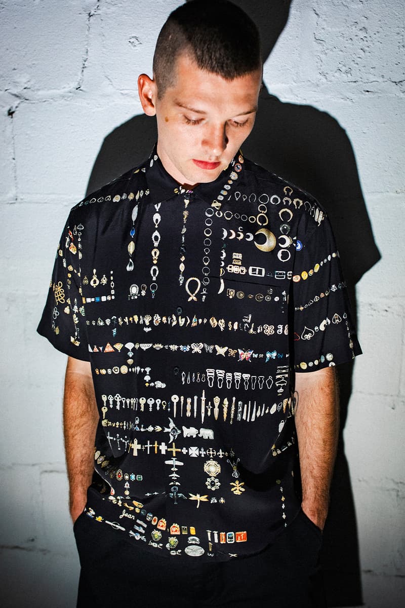 Homme avec chemise motifs bijoux, pose devant mur blanc.