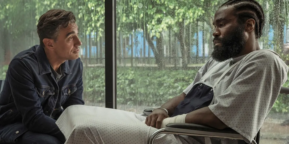 Deux hommes parlent, un en fauteuil roulant, à l'intérieur avec pluie dehors.