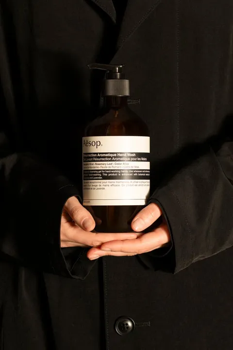 Flacon de savon pour les mains Aesop tenu par une personne vêtue de noir.