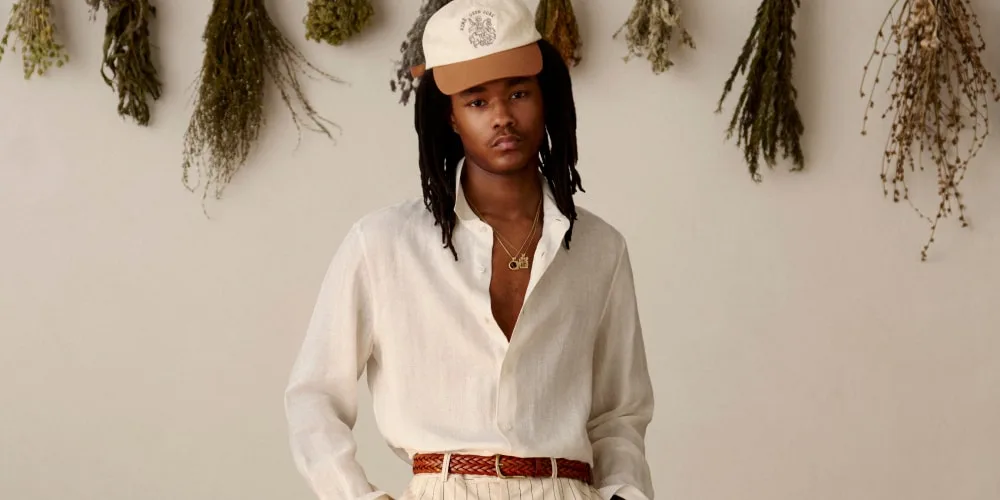 Homme en chemise blanche et casquette devant plantes séchées.