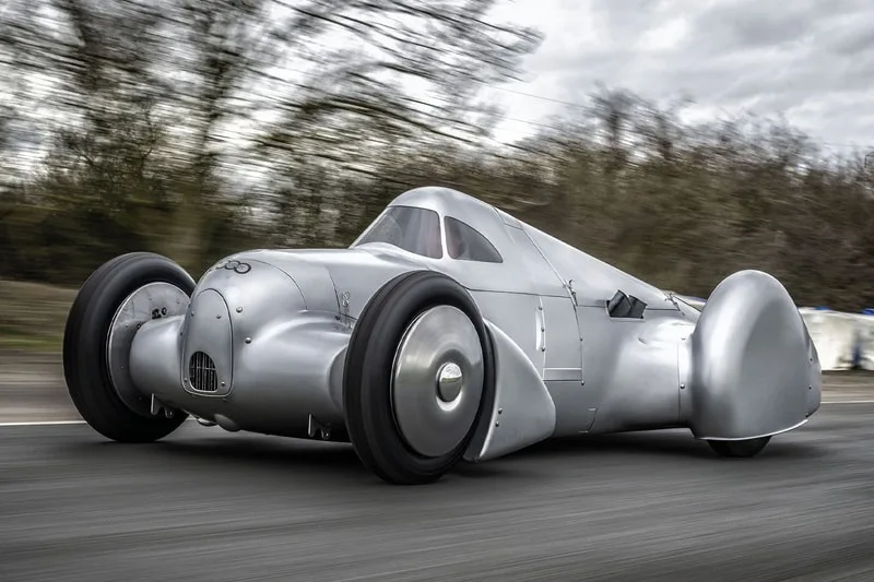 Voiture ancienne argentée sur route, design aérodynamique.