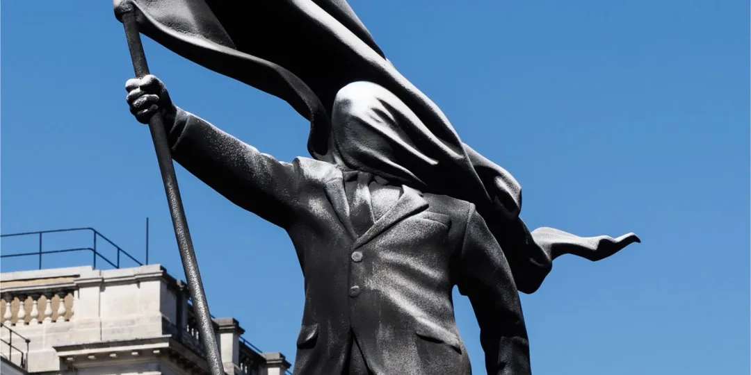 Statue de bronze avec un drapeau flottant, visage couvert, ciel bleu.