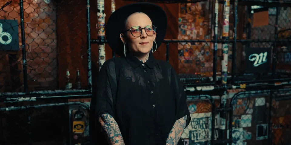 Personne tatouée avec lunettes et chapeau noir devant une clôture métallique.