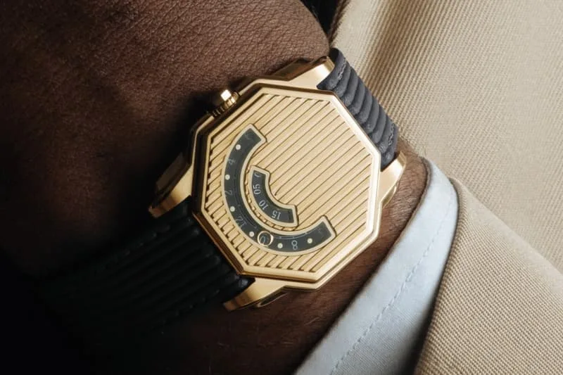Montre dorée hexagonale avec bracelet noir sur poignet habillé.