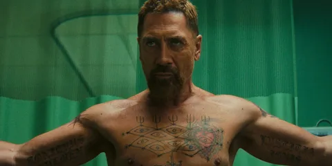 Homme tatoué au torse nu avec rideau vert en arrière-plan.