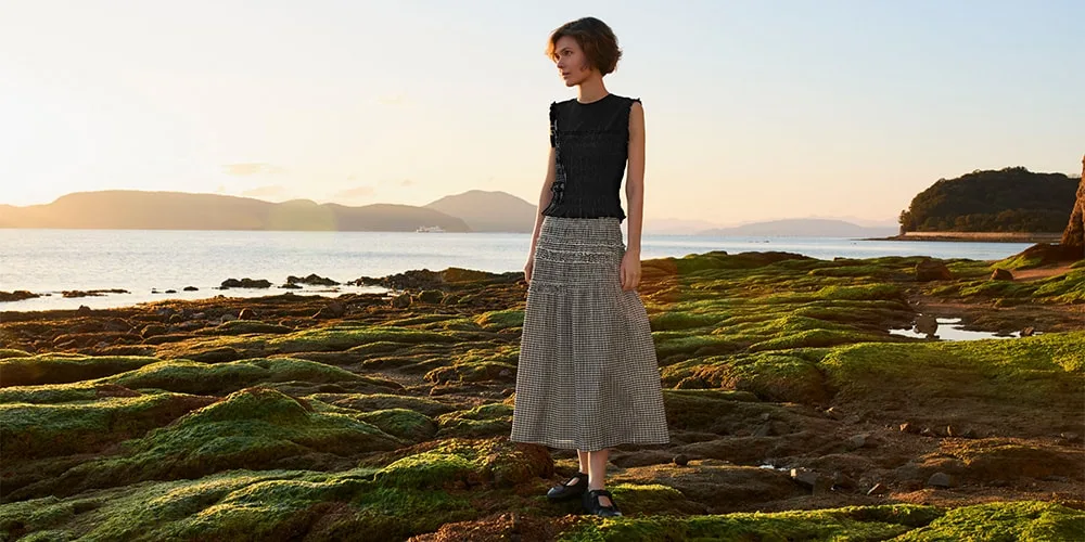 Femme debout sur un rivage rocheux au coucher du soleil, avec une jupe longue grise.