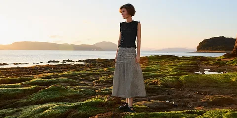 Femme debout sur un rivage rocheux au coucher du soleil, avec une jupe longue grise.