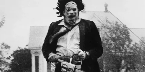 Personnage masqué avec tronçonneuse devant une maison, scène inquiétante.