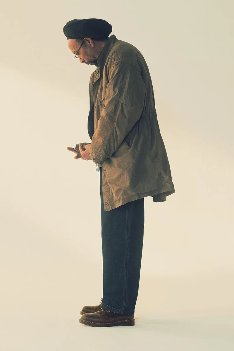 Homme avec manteau marron, pantalon noir, et lunettes, sur fond neutre.