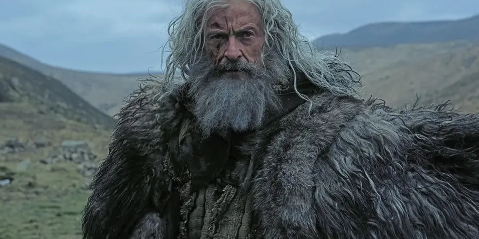 Homme barbu au manteau de fourrure dans un paysage montagneux.