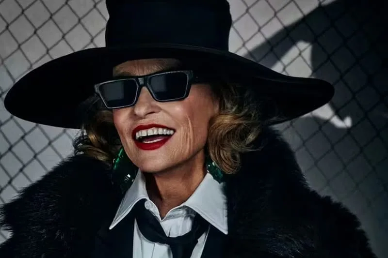 Femme portant un chapeau noir et des lunettes de soleil, souriante et élégante.