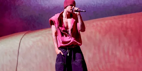 Homme chante sur scène en bonnet rouge, veste sans manches et pantalon large.