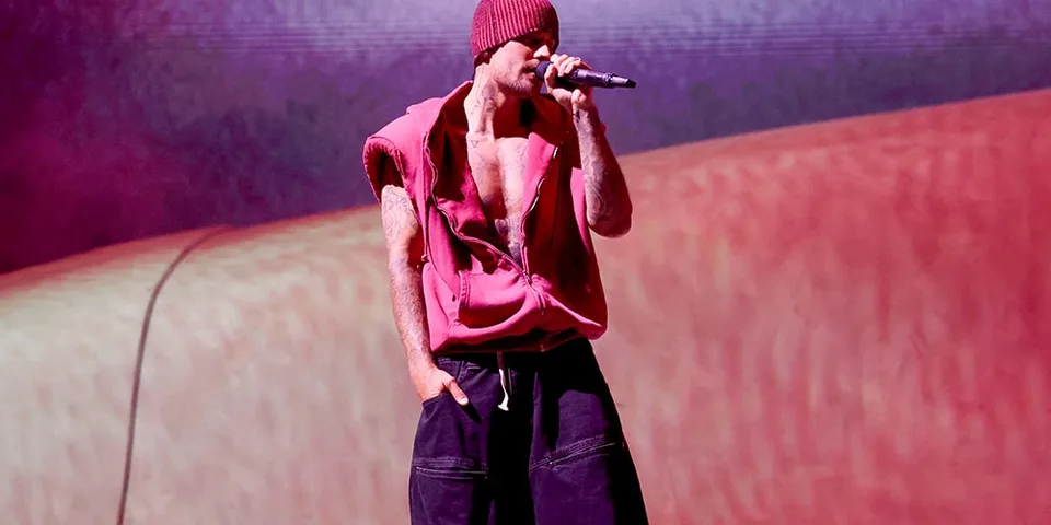 Homme chante sur scène en bonnet rouge, veste sans manches et pantalon large.