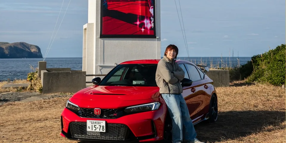 Homme souriant appuyé sur une voiture rouge près de la mer.