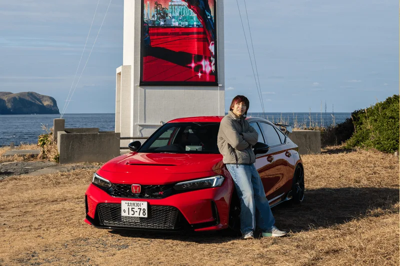 Homme posant avec une voiture rouge près de la mer, panneau en arrière-plan.