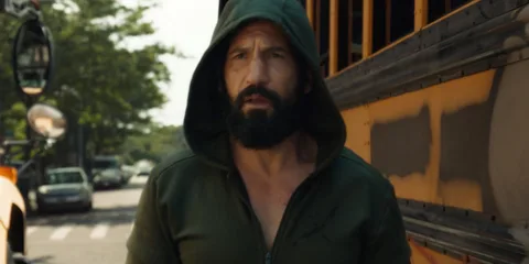 Homme barbu en sweat à capuche devant un bus scolaire jaune.