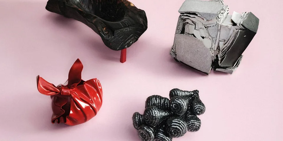 Quatre sculptures abstraites sur fond rose, dont un sac rouge et une boîte métallique argentée.