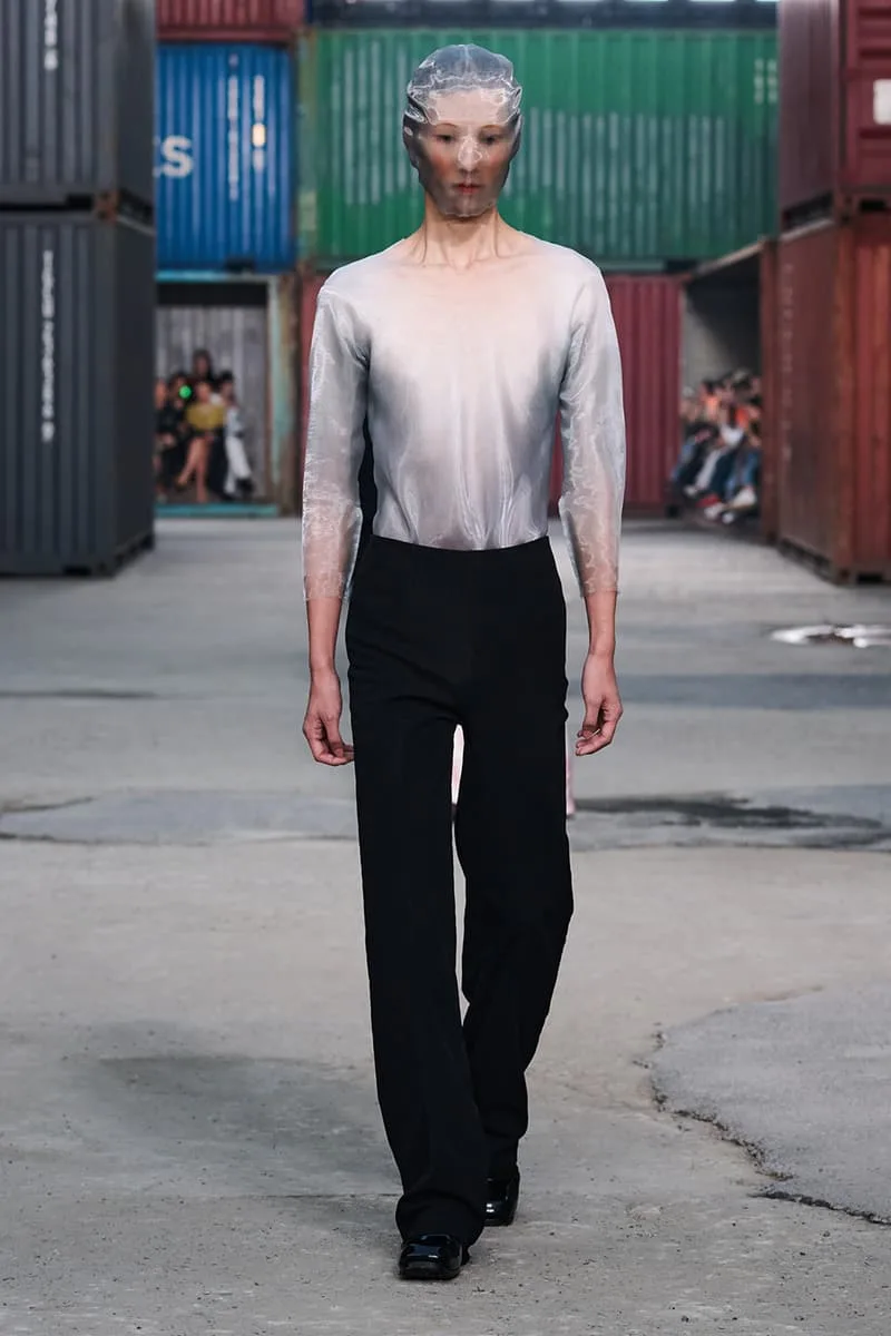 Modèle défile, haut transparent argenté, pantalon noir, décor de conteneurs colorés.