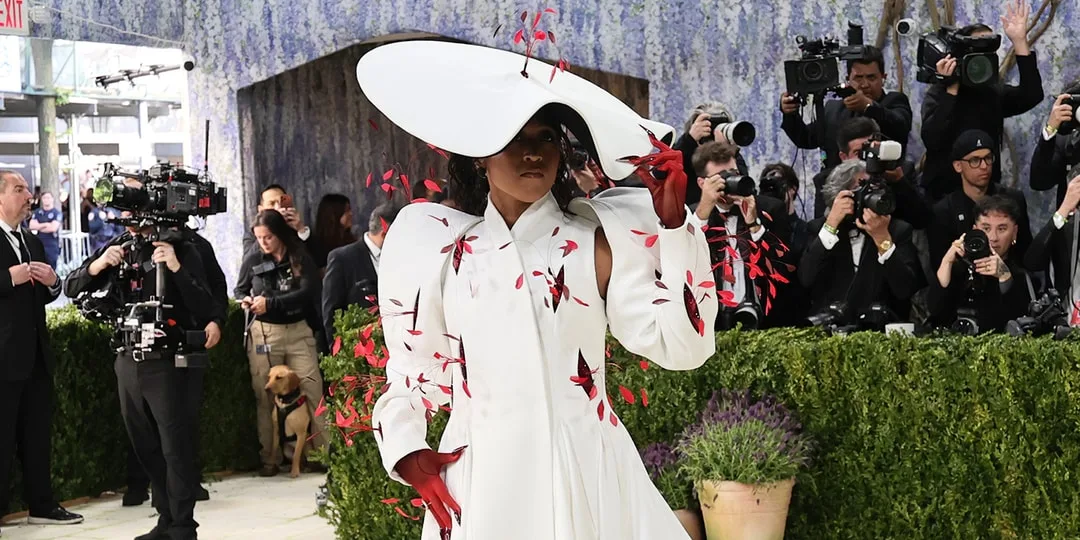 Personne en costume blanc avec des motifs rouges au milieu des photographes et des plantes.