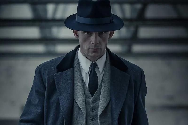 Homme en manteau et chapeau sombre, fond industriel.