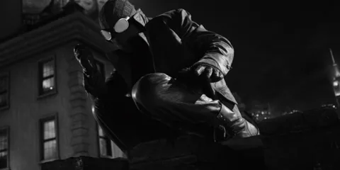 Un personnage masqué en noir et blanc accroupi sur un toit la nuit.
