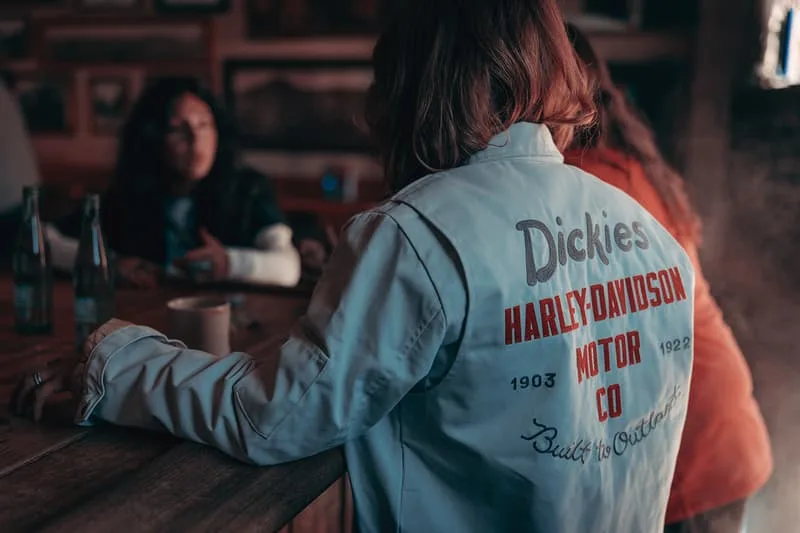 Personne portant une veste Dickies Harley-Davidson, assise à une table avec des amis.