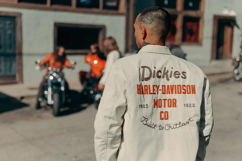 Homme en veste Dickies devant motos et bâtiment urbain.
