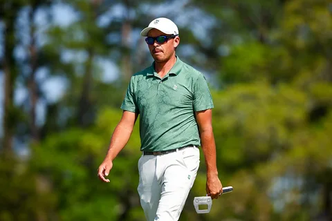 Homme marchant sur un parcours de golf, portant des lunettes de soleil et une casquette blanche.