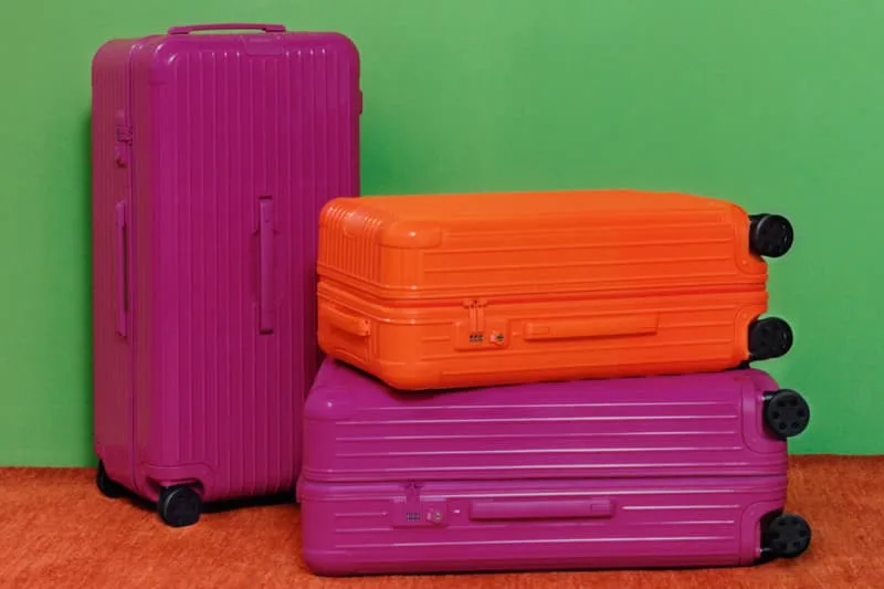 Trois valises colorées empilées, deux violettes et une orange, sur fond vert.