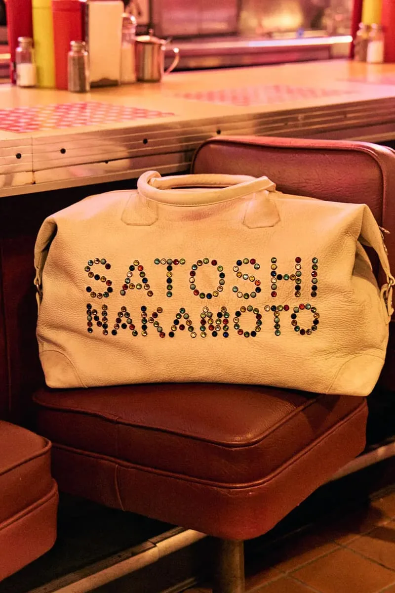 Sac beige avec texte Satoshi Nakamoto en perles, posé sur une chaise rouge dans un café rétro.