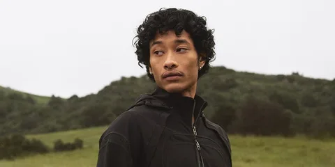 Homme en veste noire devant paysage de collines verdoyantes.