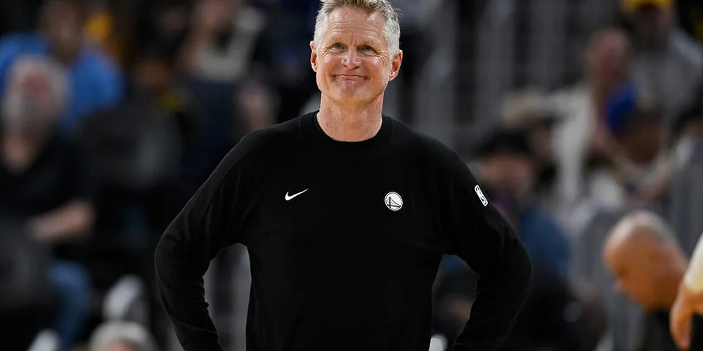 Homme souriant en pull noir avec logo sportif sur le devant.