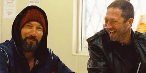 Deux hommes souriants assis ensemble, l'un avec une veste en cuir.