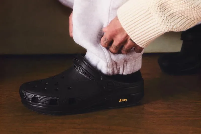 Personne portant des Crocs noires avec semelles Vibram sur un sol en bois.