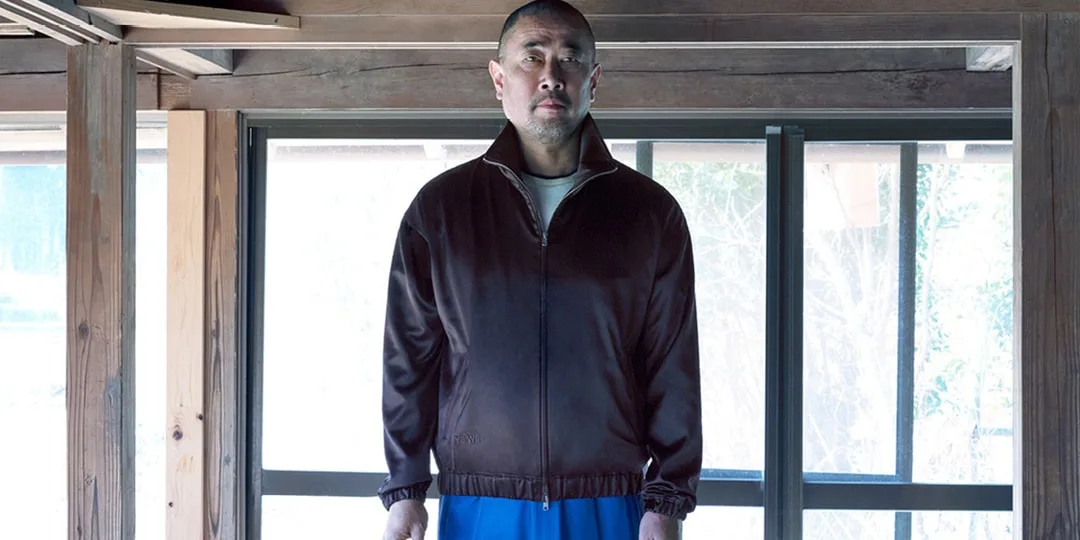 Homme en veste sombre et pantalon bleu devant fenêtre en bois.