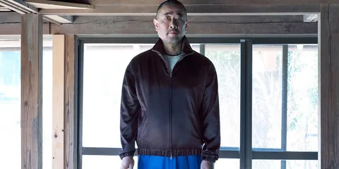 Homme en veste sombre et pantalon bleu devant fenêtre en bois.